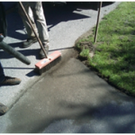 A man with a wooden broom sweeps a freshly repaired asphalt driveway